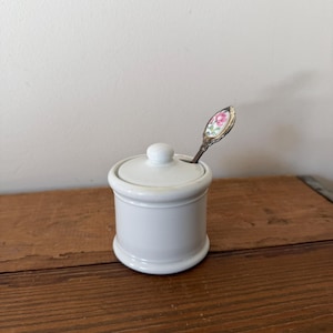 Peut inclure: Un sucrier en céramique blanche avec un couvercle et un petit bouton. Une cuillère décorative avec un motif floral est placée à l'intérieur du bol. Le bol est sur une surface en bois.