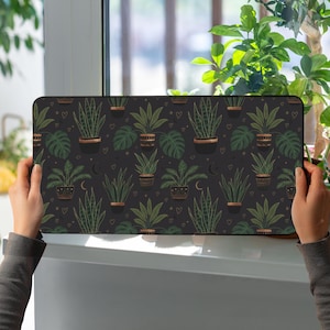 Puede incluir: Alfombrilla de ratón rectangular con un fondo gris oscuro y un patrón repetido de plantas en macetas. Las plantas están en macetas negras con detalles dorados, y el diseño incluye pequeños corazones, estrellas y lunas crecientes.