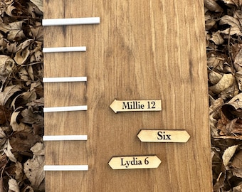 May include: A wooden growth chart with white markers and three wooden name tags. The name tags are shaped like arrows and have the names Millie 12, Six, and Lydia 6.