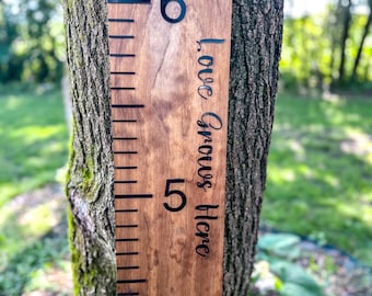 May include: A wooden growth chart with black numbers and markings, measuring up to 6 feet. The text "Love Grows Here" is written vertically on the right side. The chart is mounted on a tree trunk, with a natural wood grain finish.