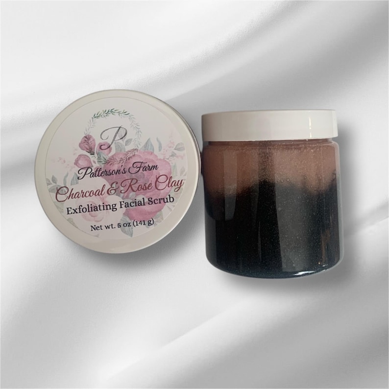 May include: A white jar with a black lid containing a charcoal and rose clay exfoliating facial scrub. The jar is labelled "Patterson's Farm Charcoal & Rose Clay Exfoliating Facial Scrub Net wt. 5 oz (141 g)".