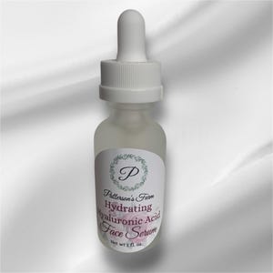 May include: A clear glass bottle with a white dropper top and a white label. The label has a green wreath with a letter "P" in the centre. The text on the label reads "Patterson's Farm Hydrating Hyaluronic Acid Face Serum Net wt 1 fl. oz."