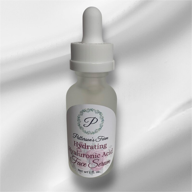 May include: A clear glass bottle with a white dropper top and a white label. The label has a green wreath with a letter "P" in the centre. The text on the label reads "Patterson's Farm Hydrating Hyaluronic Acid Face Serum Net wt 1 fl. oz."