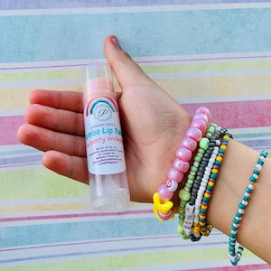 May include: A pink and white lip balm tube with a rainbow design and the text "patterson's fumy jumbo lip balm strawberry milkshake" on the label. The tube is held in a hand with several colorful beaded bracelets on the wrist.