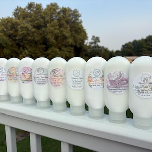May include: A row of ten white plastic squeeze bottles containing goat milk body cream, each featuring a circular label with the Patterson's Farm logo and specific scent names including Cocoa Butter Cashmere, Hot Cocoa, and Natural.