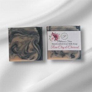 May include: Two bars of handcrafted goat milk soap with a swirl of black and light brown. The soap is labelled "Patterson's Farm Handcrafted Goat Milk Soap Rose Clay & Charcoal".