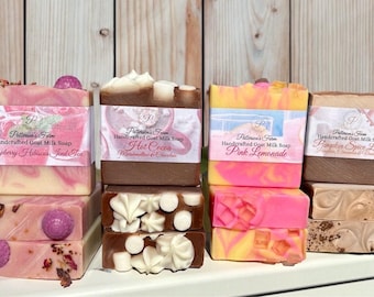 May include: A selection of handcrafted goat milk soaps, featuring scents like Raspberry Hibiscus, Hot Cocoa, Pink Lemonade, and Pumpkin Spice Latte. Each soap is decorated and labelled with its scent.