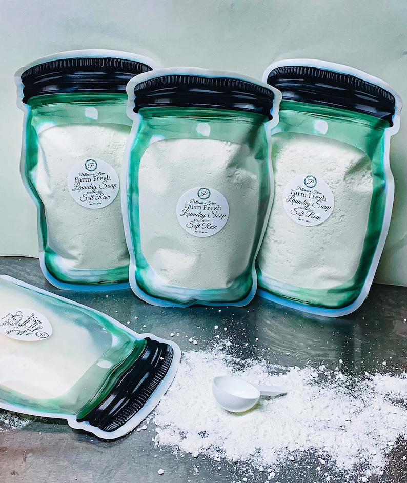 May include: Three clear plastic bags with a green jar design filled with white laundry detergent powder. The bags have a label that reads "Farm Fresh Laundry Soap Soft Rain". A white scoop is on a table with a pile of white laundry detergent powder.