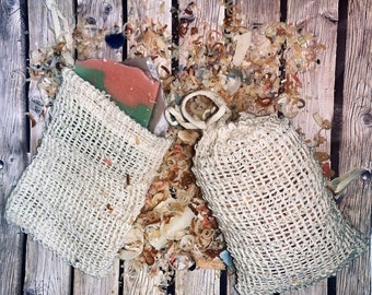 May include: Two beige mesh bags with a pink and green bar of soap inside one of the bags. The bags are on a wooden surface with wood shavings scattered around them.