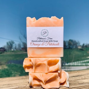 May include: Three bars of orange goat milk soap stacked on top of each other. The top bar has a white label that reads "Patterson's Farm Handcrafted Goat Milk Soap Orange & Patchouli".