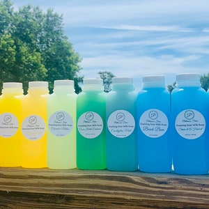 May include: A row of 15 clear plastic bottles filled with colorful liquid soap. The bottles are labeled with "Whimsical Tree" and "Soothing Goat Milk Soap". The soap colors are: Cherry Blood Orange, Pink Grapefruit,  Tangerine, Peach, Lemon, Pineapple, Green Tea, Cucumber, Mint, Ocean Breeze, Beach Plum, Spring Ocean, Lavender, and Natural.