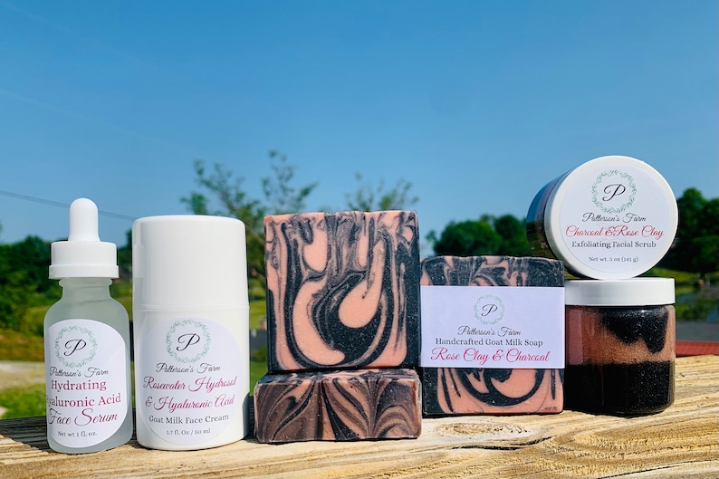 May include: A set of skincare products featuring rose clay and charcoal. The set includes a face serum, face cream, soap, and a facial scrub. The products are packaged in white containers with black and pink labels.