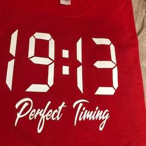 May include: Red t-shirt with white text that reads "19:13 Perfect Timing".