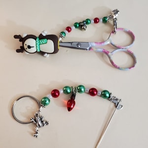 Può includere: Forbici decorative con un ciondolo a forma di renna e una catena di perline rosse, verdi e bianche. Sono inclusi anche un ciondolo a forma di rana e una lampadina rossa.