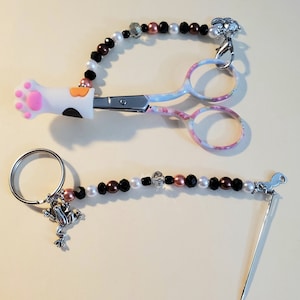 Può includere: Forbici decorative con un motivo floreale e un manico a forma di zampa di gatto, e un portaghiaccio. Entrambi sono adornati con fili di perline nere, bianche e rosa, con ciondoli argentati. Le forbici hanno una chiusura argentata e il portaghiaccio ha un ciondolo a forma di rana.