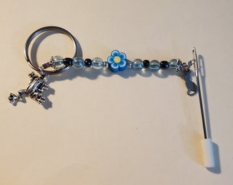 Könnte beinhalten: Ein Fädelhilfe mit Perlen, einem silbernen Frosch-Anhänger und einer blauen Blumenperle. Der Einfädler hat eine silberne Nadel und einen weißen Schaumstoffgriff. Die Perlen sind klar und dunkelblau.