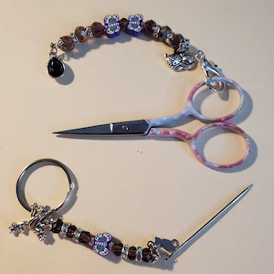 May include: A set of craft tools including a pair of small scissors with floral handles, a needle, and a beaded charm with a black teardrop, butterfly, and cat accents. The scissors are approximately 4 inches long.