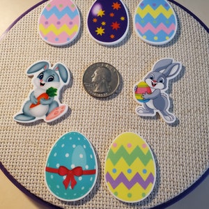 May include: A selection of Easter-themed decorations, featuring patterned egg shapes and two cartoon bunnies. A U.S. quarter is included for size reference. The decorations are arranged on a neutral-coloured fabric.