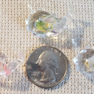 May include: Three clear, glass teapot figurines with colorful inclusions. One quarter coin is next to the teapots for size comparison. The teapots are on a textured, tan surface.