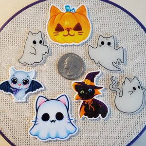 May include: A selection of Halloween-themed acrylic charms. The charms include a pumpkin cat, a black cat in a witch hat, ghosts, and a bat. The charms are in orange, black, white, and blue. A coin is included for size reference.