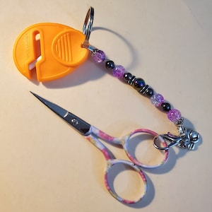 May include: An orange Fiskars thread cutter and small floral scissors with a beaded charm. The scissors have a colorful pink, yellow, and white pattern on the handles. The charm features purple and black beads.