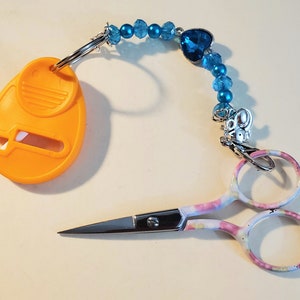 May include: A pair of small, decorative scissors with a colorful floral pattern on the handles, connected to a key ring and an orange thread cutter. The scissors are attached to a beaded blue charm with a heart-shaped jewel.