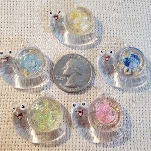 May include: Five clear resin snail pendants with smiling faces. Each snail's shell contains colorful elements: yellow, blue, green, pink, and blue with gold flakes. A US quarter is included for size reference.