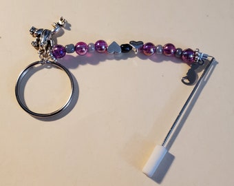 Könnte beinhalten: Ein silberfarbener Schlüsselanhänger mit einem Frosch-Anhänger, einem Perlenabschnitt mit lila und grauen Perlen und einer Nadel mit einem weißen Stopper. Der Perlenabschnitt enthält herzförmige Perlen. Der Schlüsselanhänger ist für Schlüssel konzipiert.