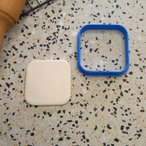 May include: A white square of fondant is placed on a speckled countertop next to a blue plastic square cookie cutter.
