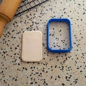 May include: A white fondant rectangle and a blue plastic cookie cutter in the shape of a rectangle on a speckled work surface.