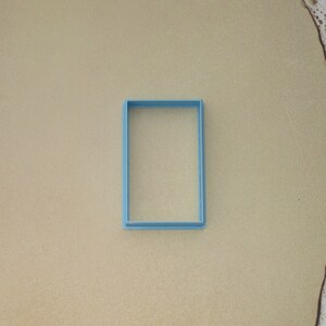 May include: A light blue plastic rectangle cookie cutter on a beige surface.