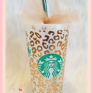 May include: A white reusable cup with a gold leopard print design and a green Starbucks logo. The cup has a clear plastic lid and a green straw.