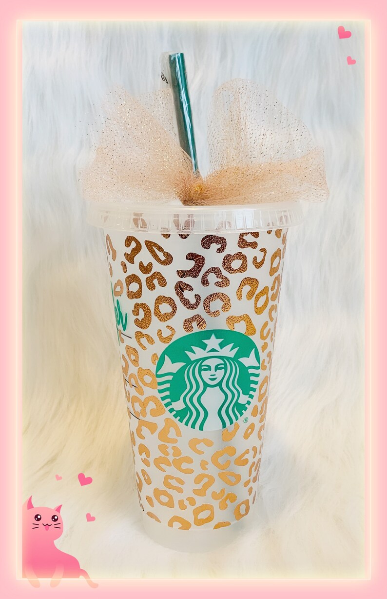 May include: A white reusable cup with a gold leopard print design and a green Starbucks logo. The cup has a clear plastic lid and a green straw.