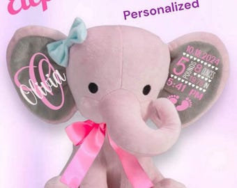 May include: A pink plush elephant with gray ears, a pink bow, and a blue bow on its head. The ears are personalized with a name, birth date, time, weight, and length. The elephant is sitting on a pink background.  The text on the ears reads "Olivia", "10.18.2024", "5 pounds 8 ounces", "19.75 in", "5:41 PM".