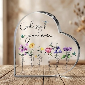 May include: A heart-shaped clear acrylic plaque featuring the text God says you are... followed by six floral illustrations with the words unique, special, lovely, precious, strong, and chosen, each paired with small hummingbird graphics.