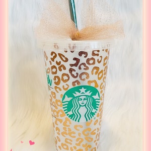 May include: A white reusable cup with a gold leopard print design and the Starbucks logo. The cup has a gold tulle bow and a green straw.