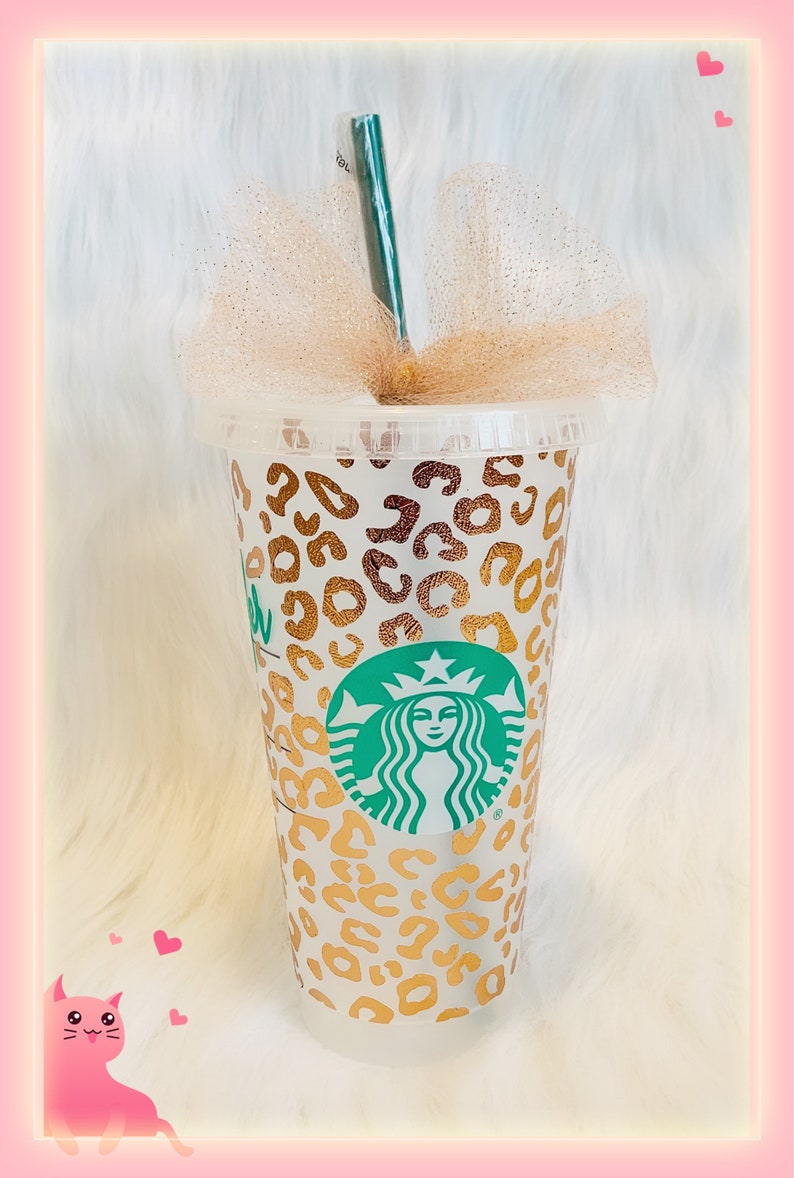 May include: A white reusable cup with a gold leopard print design and the Starbucks logo. The cup has a gold tulle bow and a green straw.