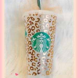 May include: A clear plastic reusable cup with a gold and white leopard print design. The cup has the Starbucks logo in green and white on the front.