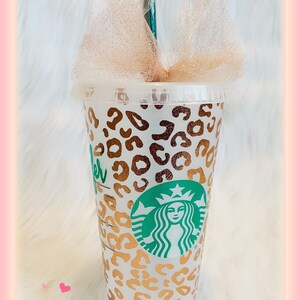 May include: A clear reusable cup with a gold leopard print design and the Starbucks logo. The cup has a straw and a gold glitter bow on top.