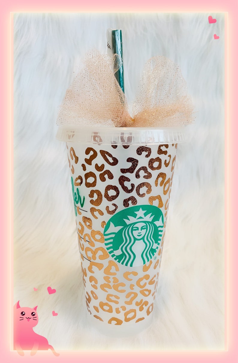 May include: A clear reusable cup with a gold leopard print design and the Starbucks logo. The cup has a straw and a gold glitter bow on top.
