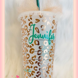 May include: A clear plastic reusable cup with a gold leopard print design and the name "Jennifer" written in teal on the front. The cup has a green straw and is sitting on a white fluffy surface.