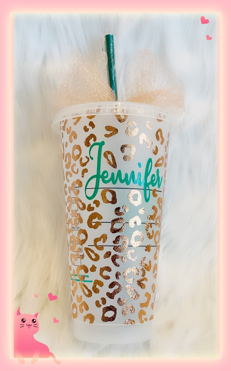 May include: A clear plastic reusable cup with a gold leopard print design and the name "Jennifer" written in teal on the front. The cup has a green straw and is sitting on a white fluffy surface.