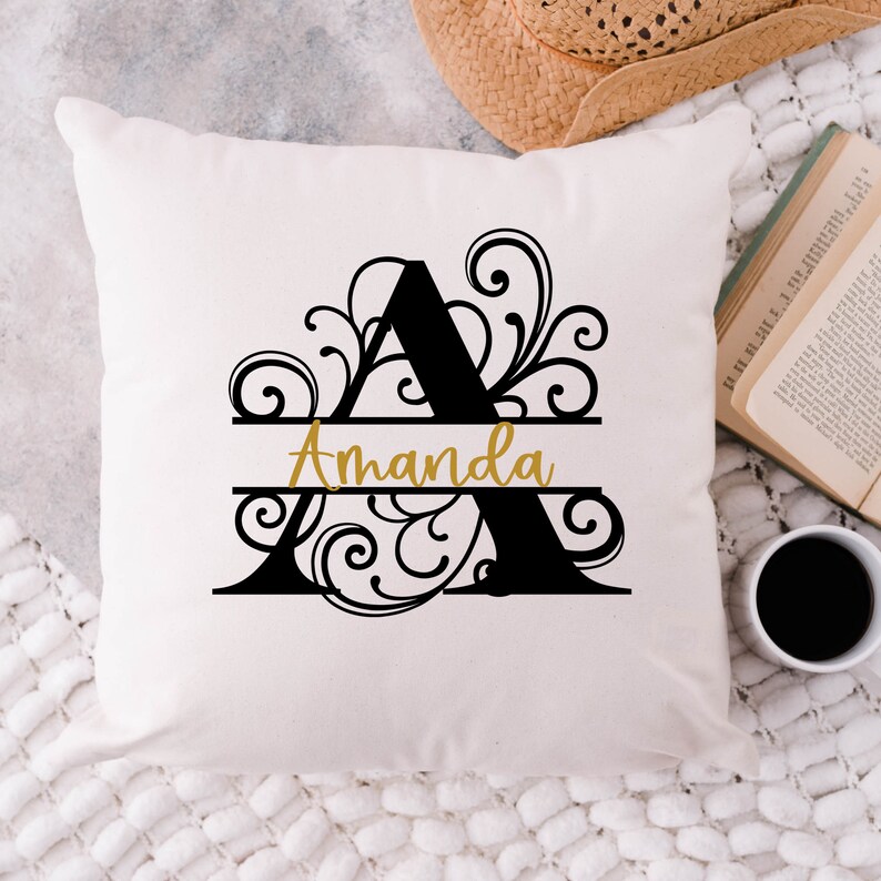 May include: Cream-colored throw pillow with a large black letter "A" and the name "Amanda" in gold script. The letter is decorated with ornate swirls. A straw hat, open book, and coffee cup are in the background.