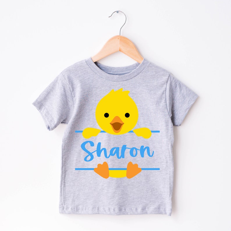 May include: A light gray t-shirt with a cartoon yellow chick design. The chick is peeking over a blue line with the name "Sharon" in blue script. The shirt is hanging on a wooden hanger.