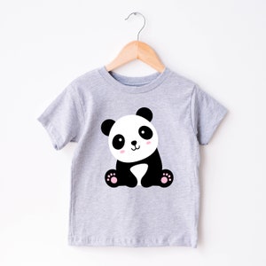 May include: A light gray t-shirt featuring a cartoon panda bear. The panda has black ears, eyes, and limbs, with a white face and body. The panda's cheeks are pink, and it has pink paw pads. The shirt is hanging on a wooden hanger.