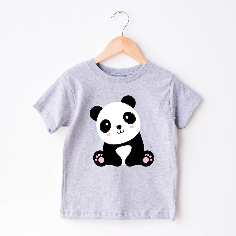 May include: A light gray t-shirt featuring a cartoon panda bear. The panda has black ears, eyes, and limbs, with a white face and body. The panda's cheeks are pink, and it has pink paw pads. The shirt is hanging on a wooden hanger.