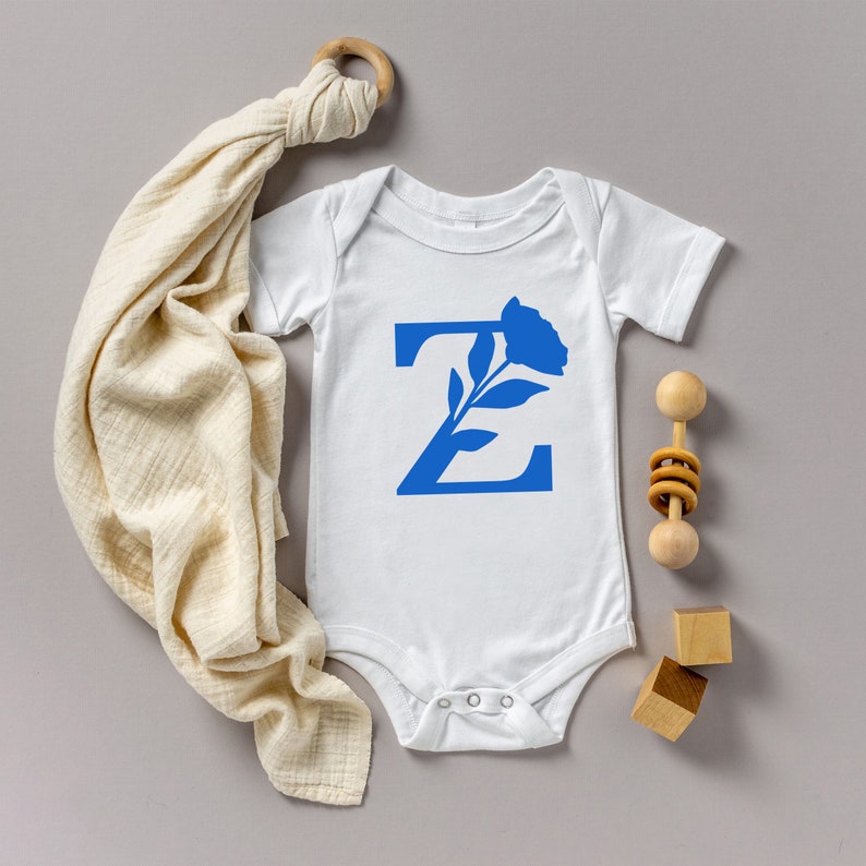 Pu&ograve; includere: Body da bambino bianco con un design floreale blu a forma di lettera "Z". Un sonaglio di legno, due piccoli blocchi di legno e una coperta color crema sono disposti attorno al body su una superficie grigia. Il body ha maniche corte e bottoni automatici sul fondo.