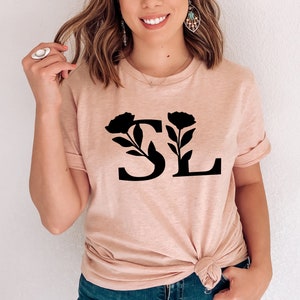 Pu&ograve; includere: Una maglietta color pesca chiaro con le lettere nere "SL" e dettagli floreali. La "S" &egrave; decorata con un design floreale a sinistra, e la "L" ha un elemento floreale simile a destra. La maglietta &egrave; annodata in vita.