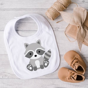 May include: A white baby bib featuring a gray cartoon raccoon illustration. The bib is surrounded by a spool of twine, a wrapped gift, and a pair of tan baby shoes. The bib is designed for infants and toddlers.