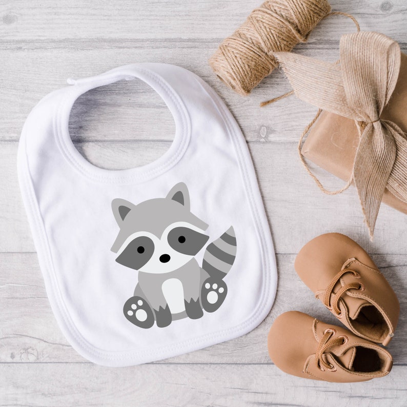 May include: A white baby bib featuring a gray cartoon raccoon illustration. The bib is surrounded by a spool of twine, a wrapped gift, and a pair of tan baby shoes. The bib is designed for infants and toddlers.